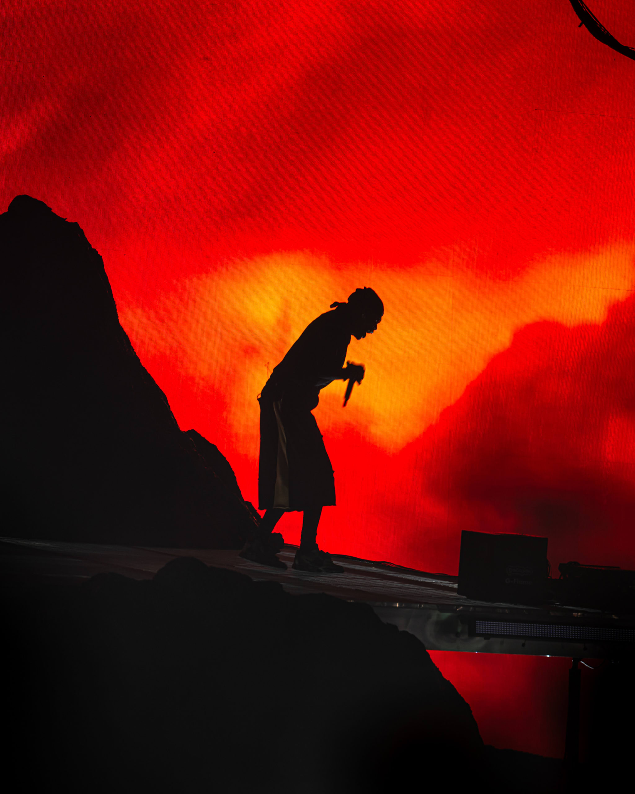 Silhouette of Travis Scot with a microphone against a vivid red and orange background, resembling a sunset or fiery scene.