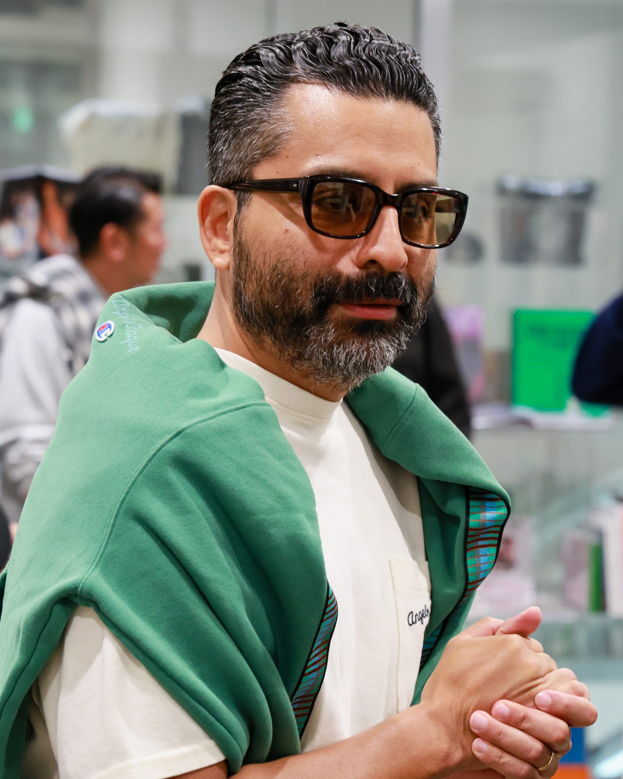 A man with sunglasses and a beard, wearing a white shirt and green sweater draped over his shoulders, standing indoors.