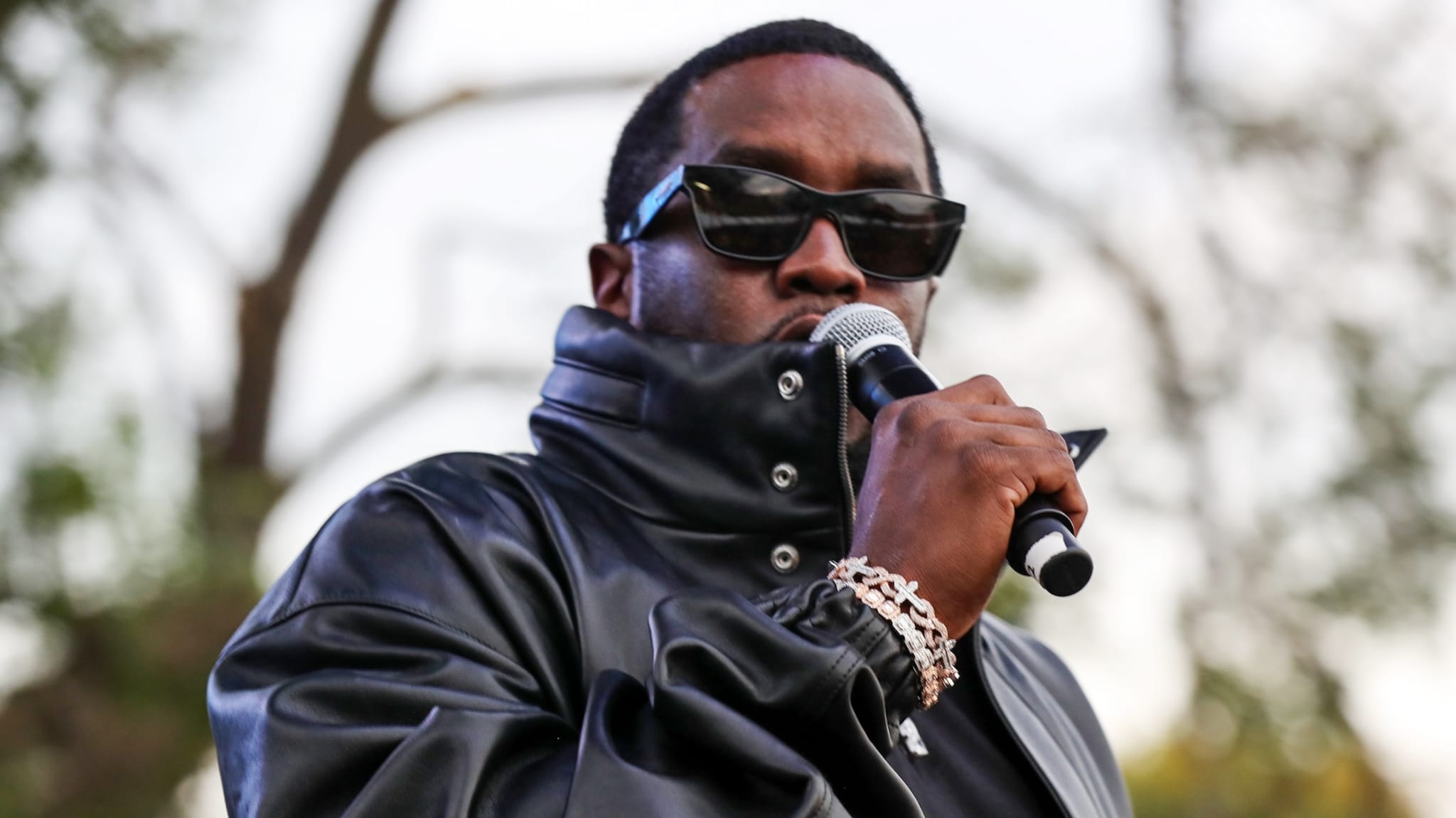 Sean "Diddy" Combs wearing black leather jacket and sunglasses while holding microphone.