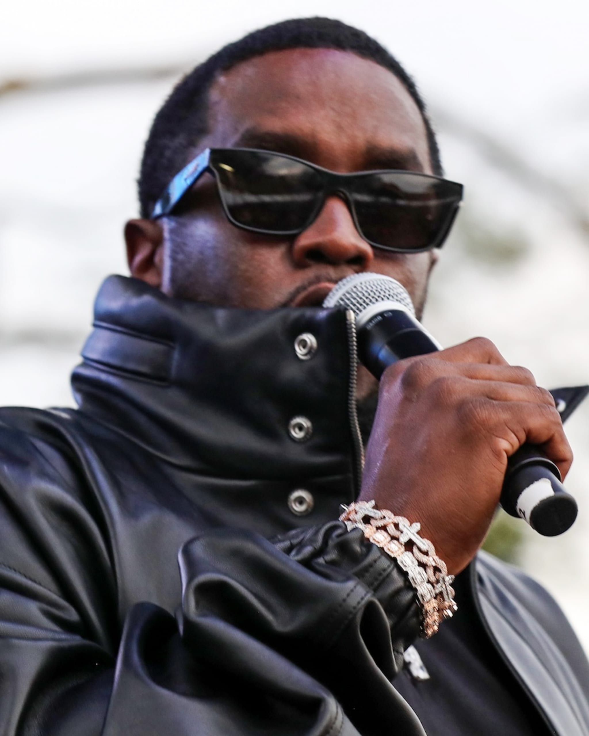 Sean "Diddy" Combs wearing black leather jacket and sunglasses while holding microphone.