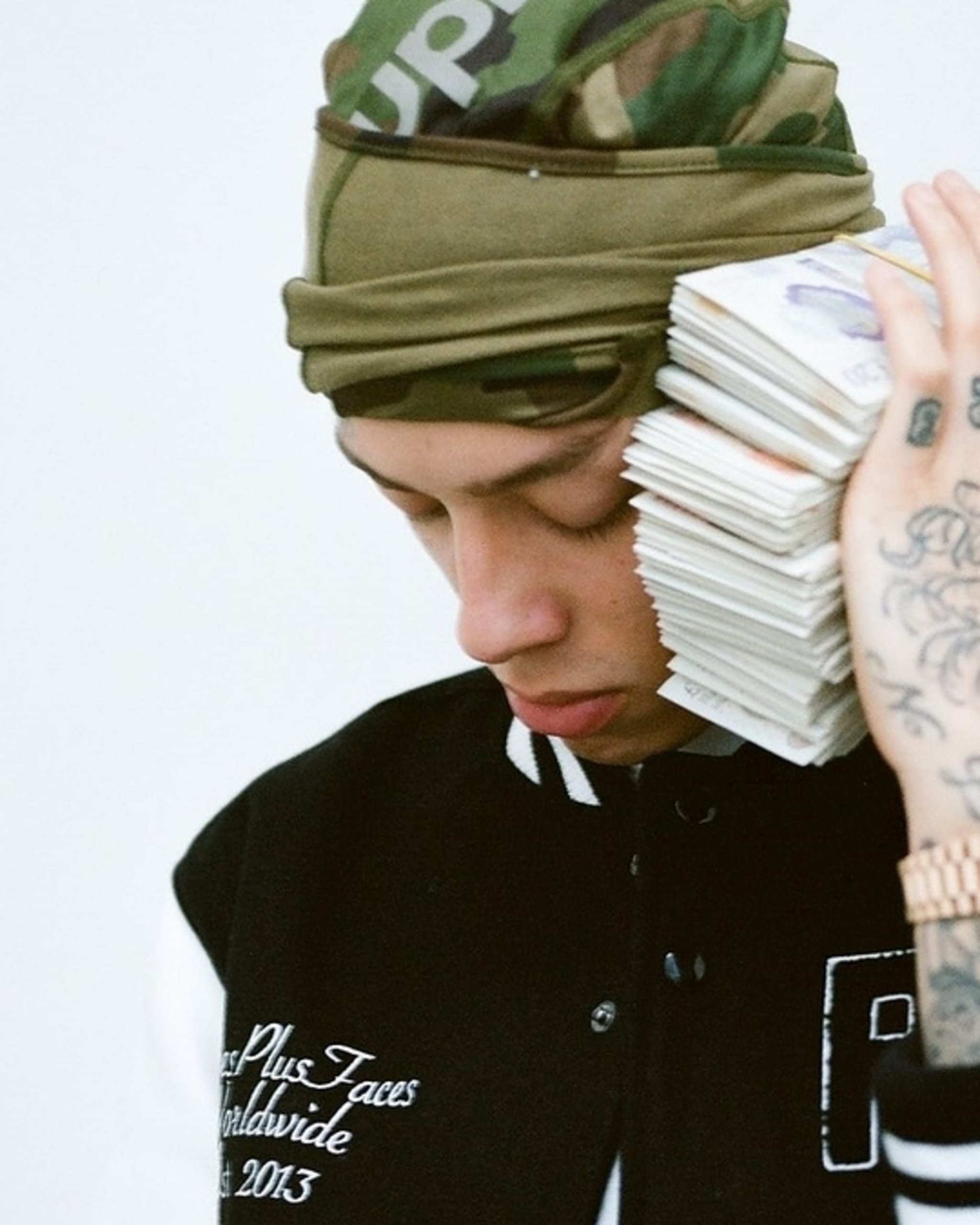 A person wearing a camo beanie and a black and white varsity jacket holds a large stack of money to their ear like a phone. Their hand has tattoos