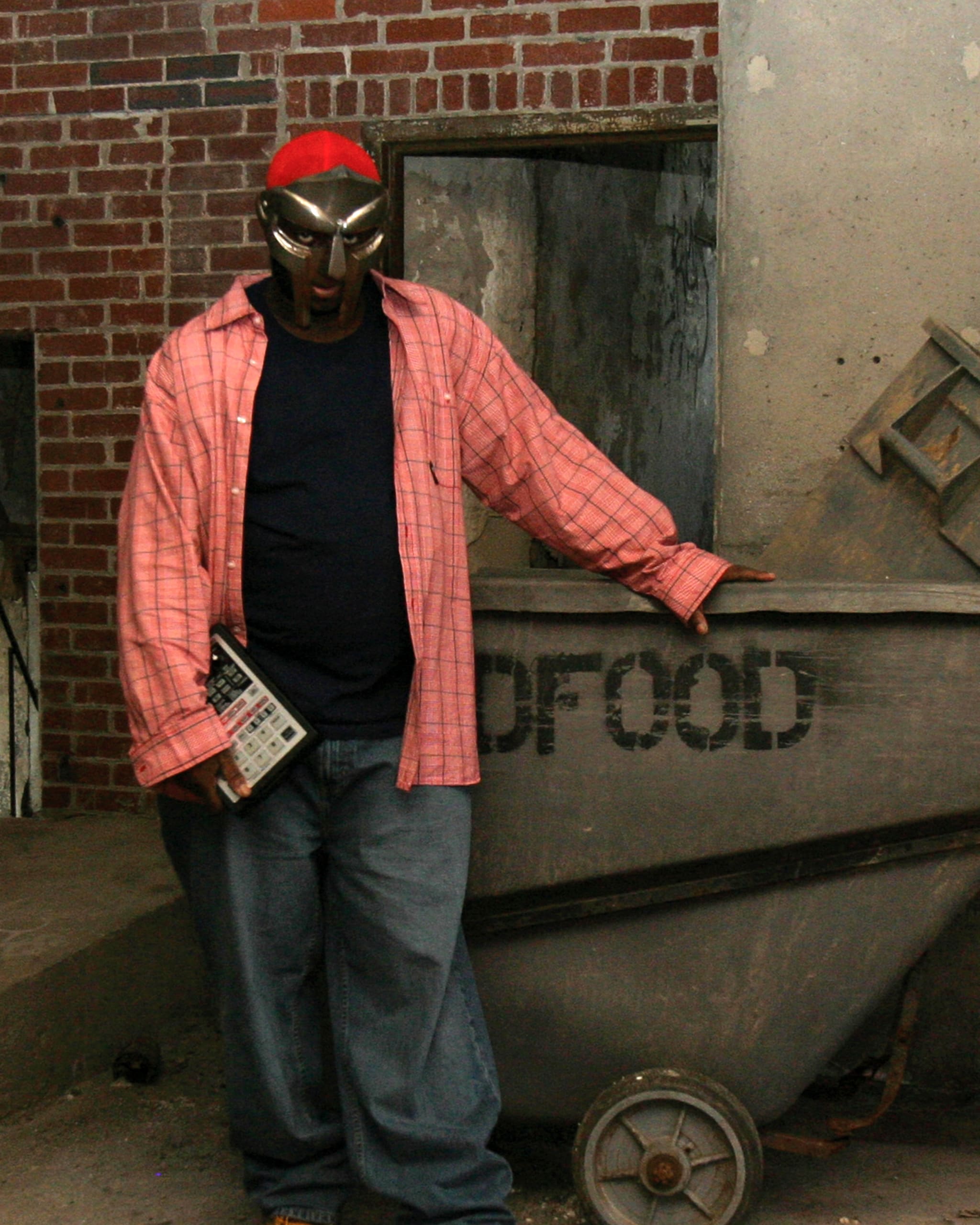 MF DOOM holding a device, stands next to a large metal container against a brick wall.