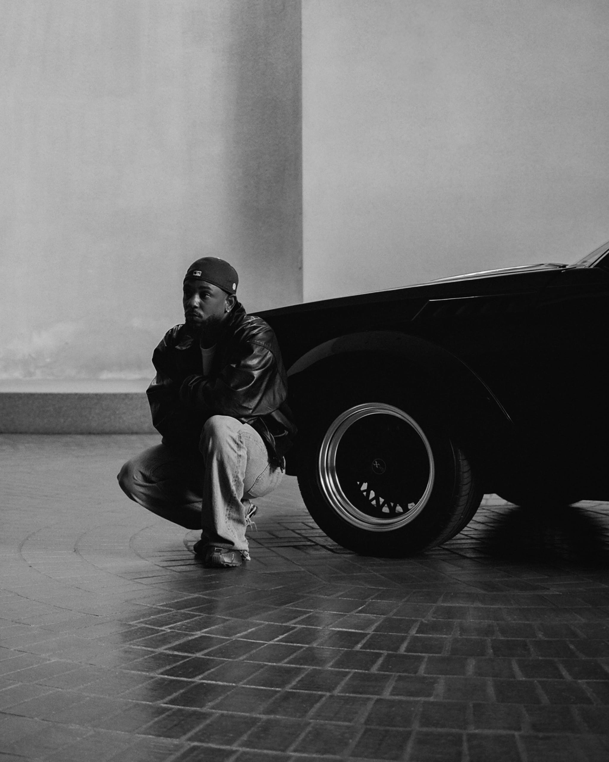 Kendrick Lamar squatting by a car in a large, empty, tiled space. The image is in black and white, creating a dramatic and moody atmosphere.