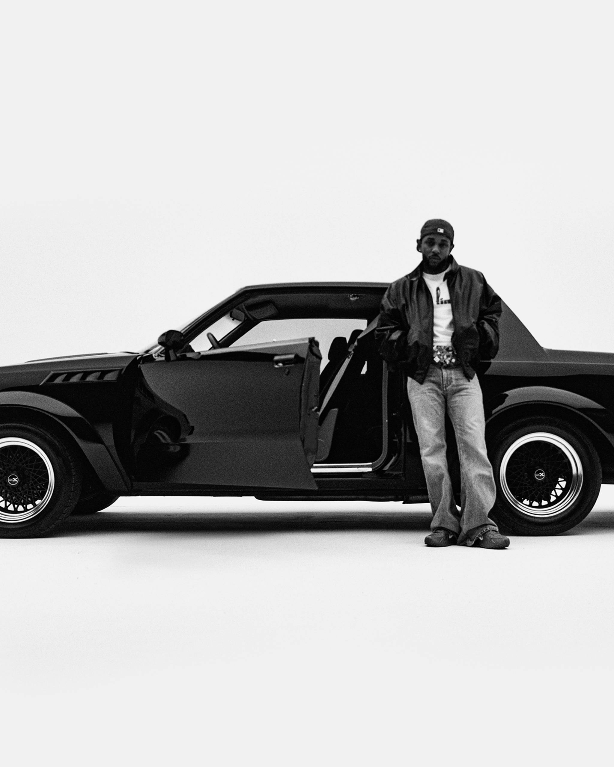 Kendrick Lamar in a leather jacket and jeans stands beside an open door of a classic black car in a minimalist setting.