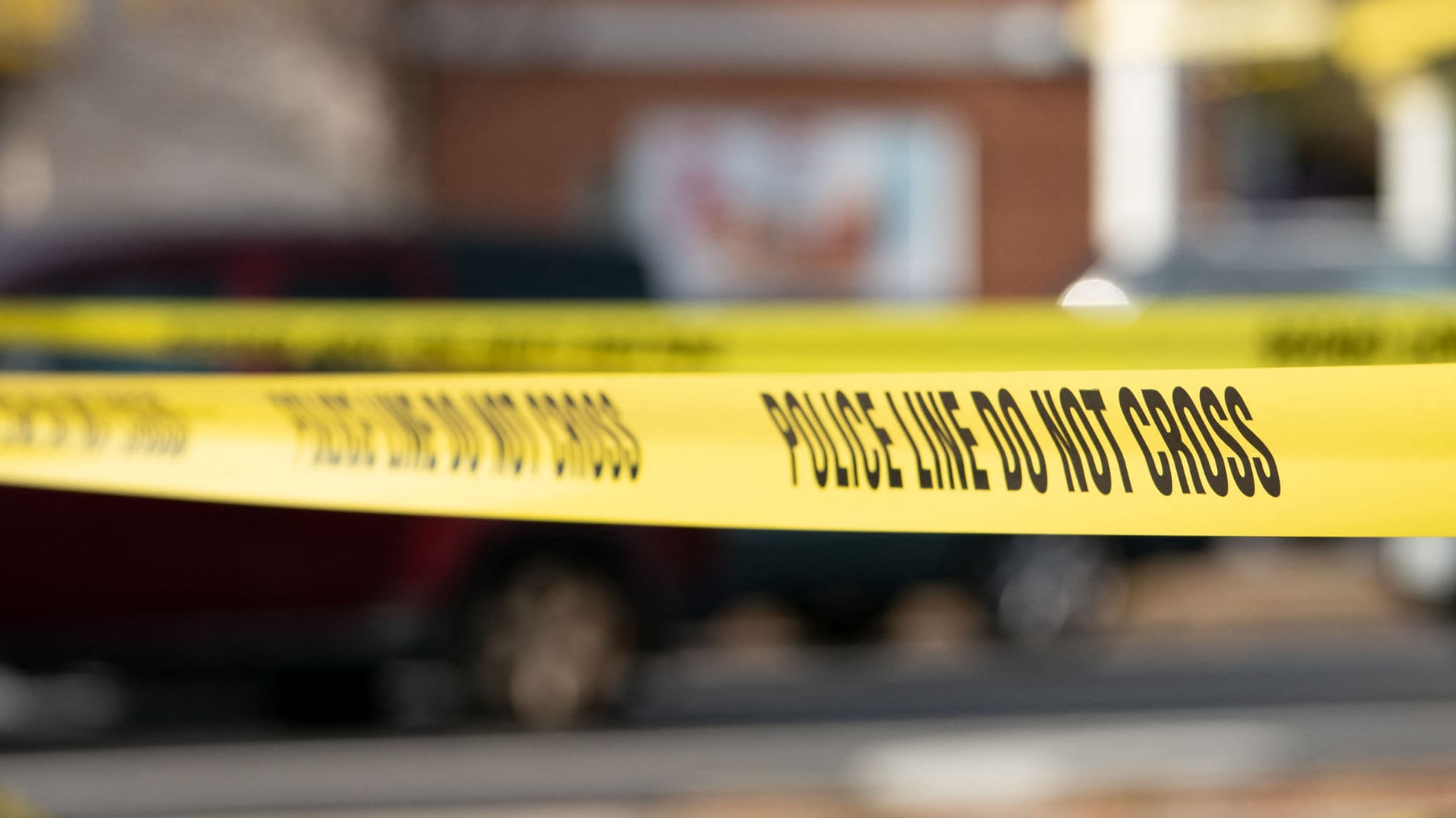 Yellow police tape with "POLICE LINE DO NOT CROSS" text, blurred background with a vehicle and building.