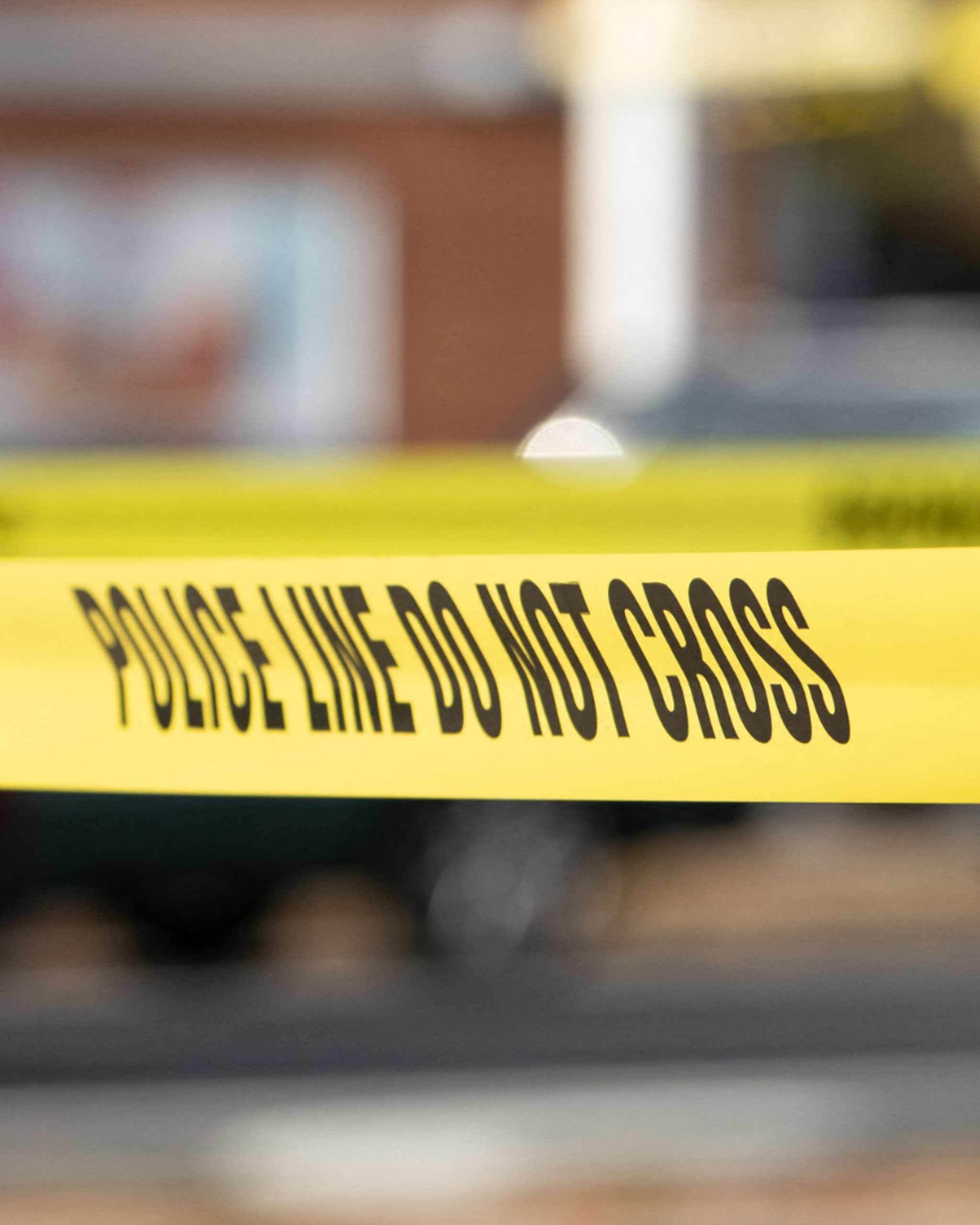 Yellow police tape with "POLICE LINE DO NOT CROSS" text, blurred background with a vehicle and building.