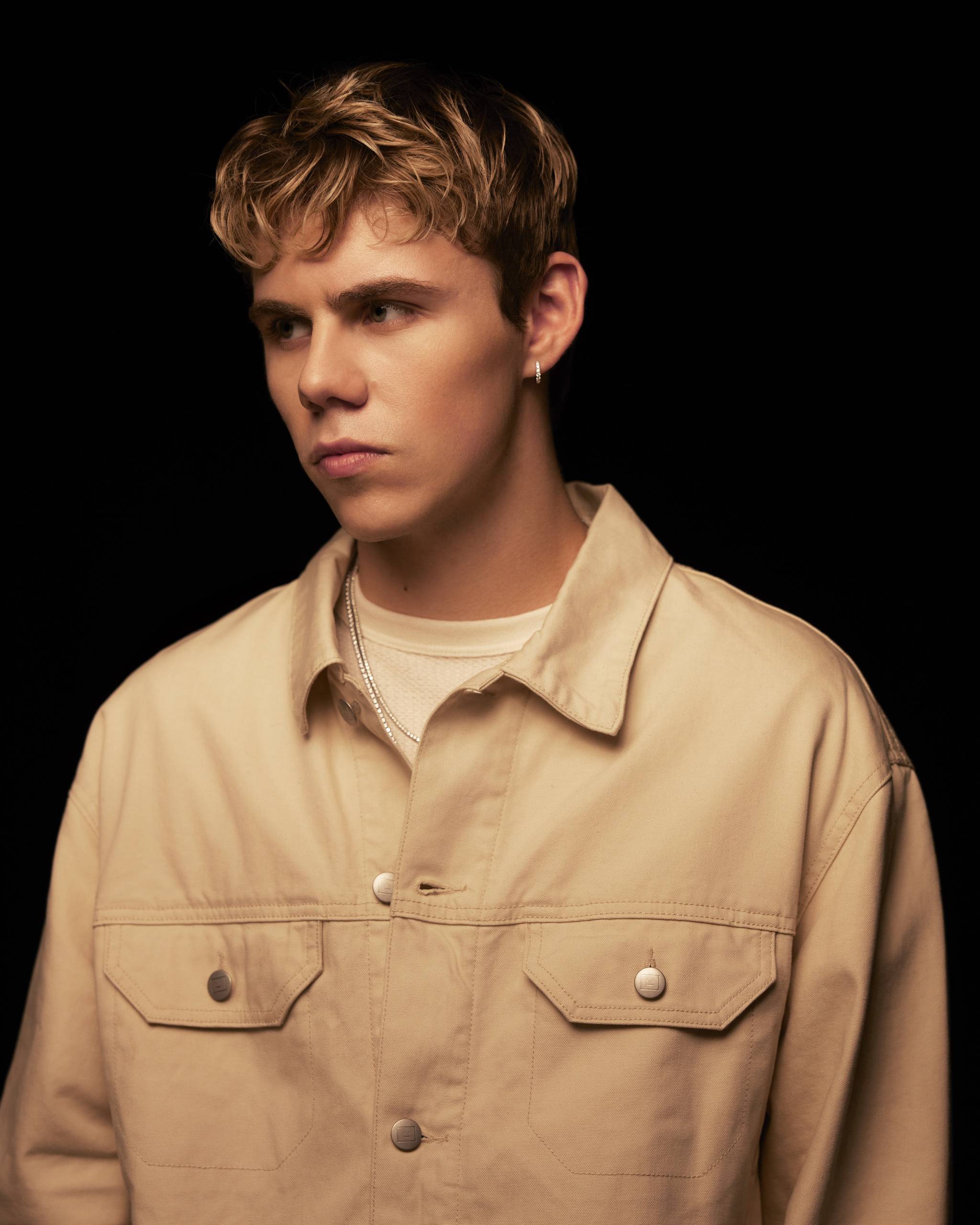 Young man with short, wavy hair wearing a beige jacket, looking off to the side against a dark background.